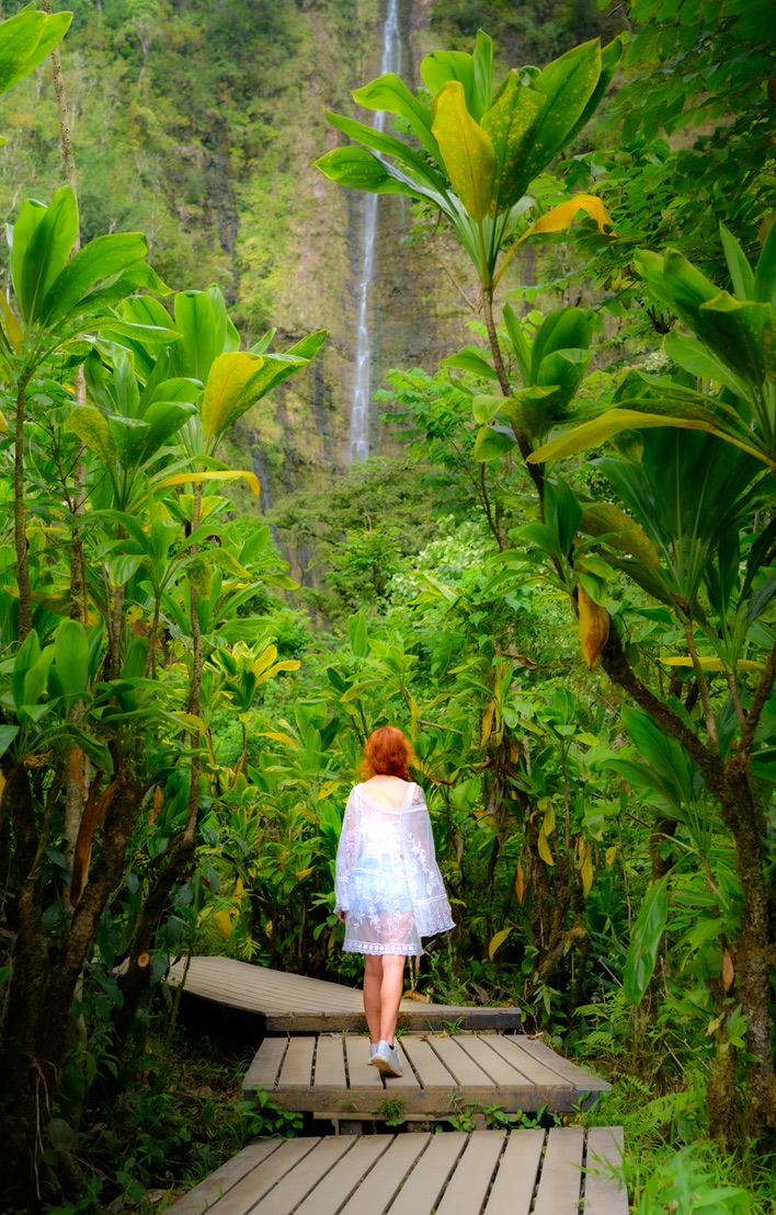 A woman walking along a boardwalk through lush foliage toward a waterfall